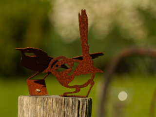 Rusty metal wren sculpture perched on a weathered wooden post with a blurred green garden background