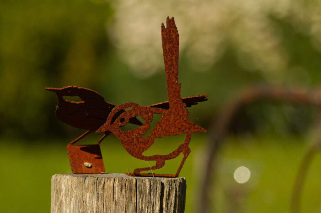 Rusty metal wren sculpture perched on a weathered wooden post with a blurred green garden background