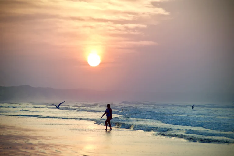 A lone swimmer walks along the shoreline at Ocean Grove during sunrise with soft waves and warm morning light.