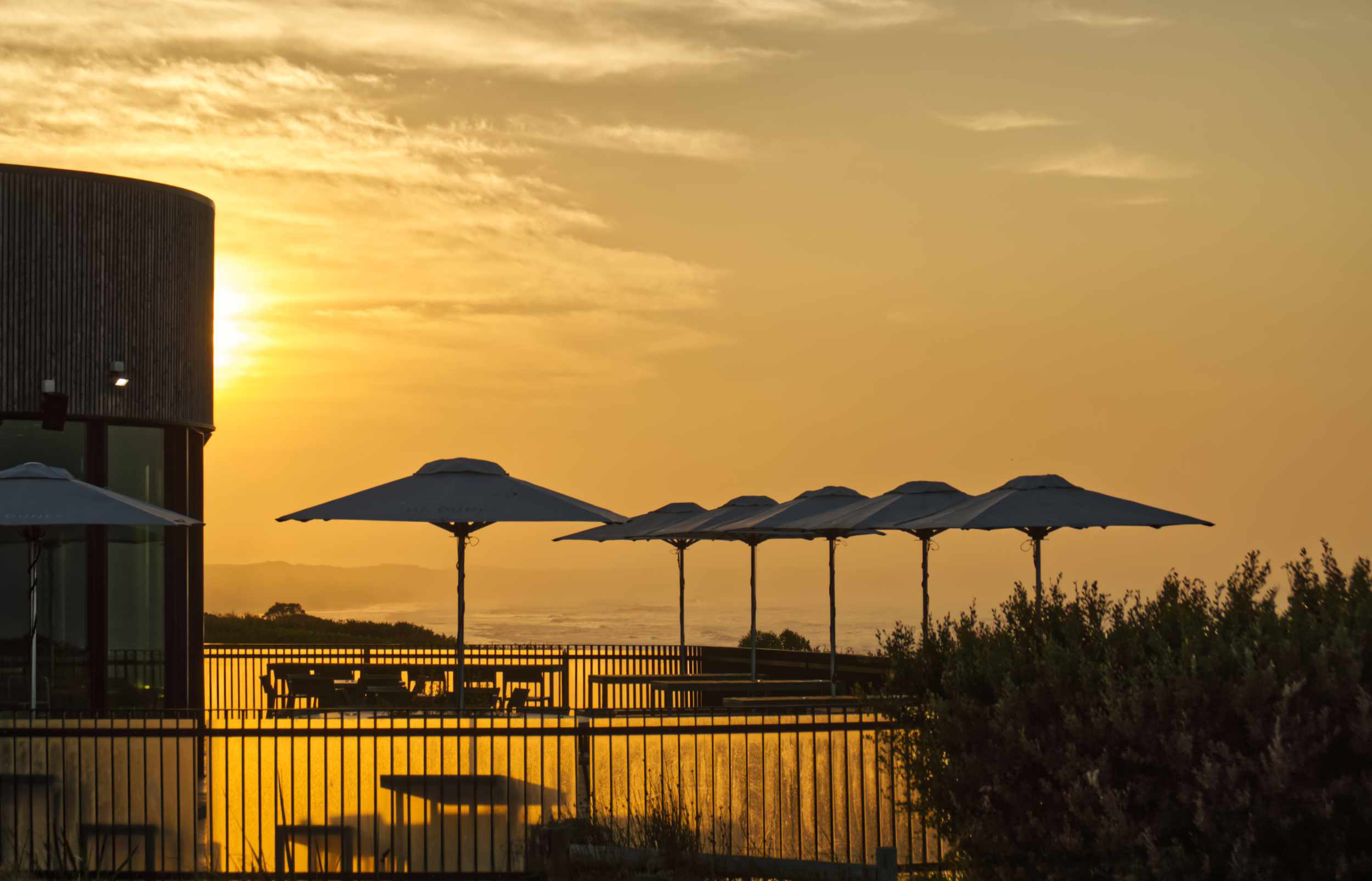 Sunrise casts golden light across a coastal café terrace with umbrellas overlooking the dunes and ocean.