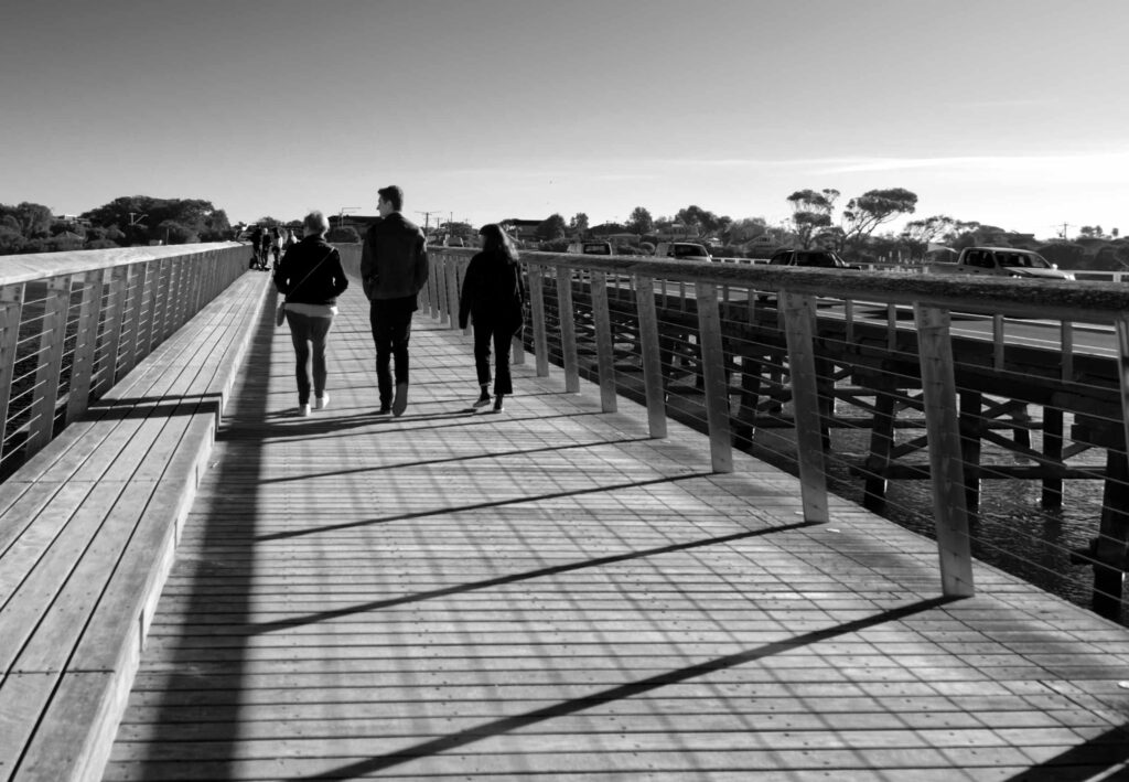 Black and white photo of people walking across the Barwon Heads Bridge over the Barwon River, Victoria, with long shadows cast by the railings.