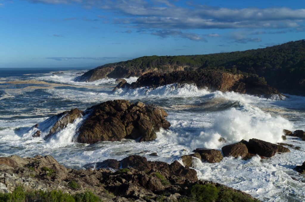 Powerful waves crash against rugged coastal rocks near Tathra under a bright blue sky.