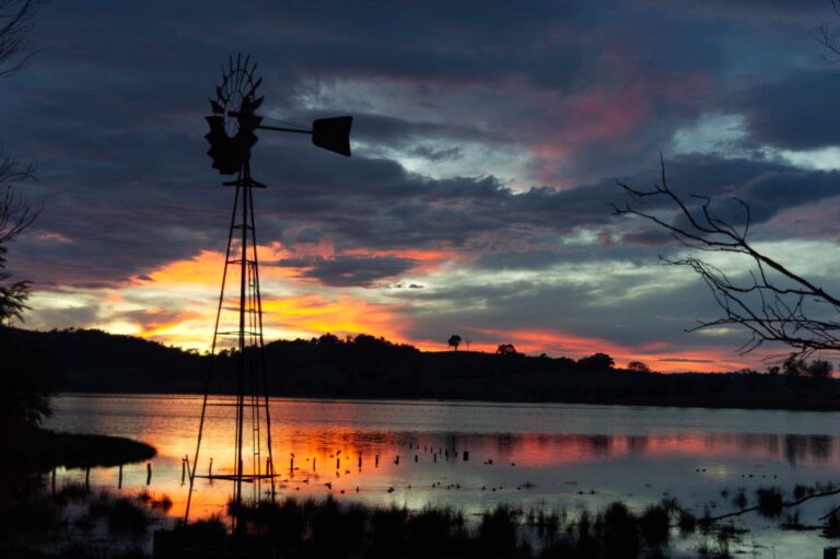 Silhouetted rural windmill standing beside calm water at sunset, with orange and pink clouds reflecting across a quiet lake under a darkening sky